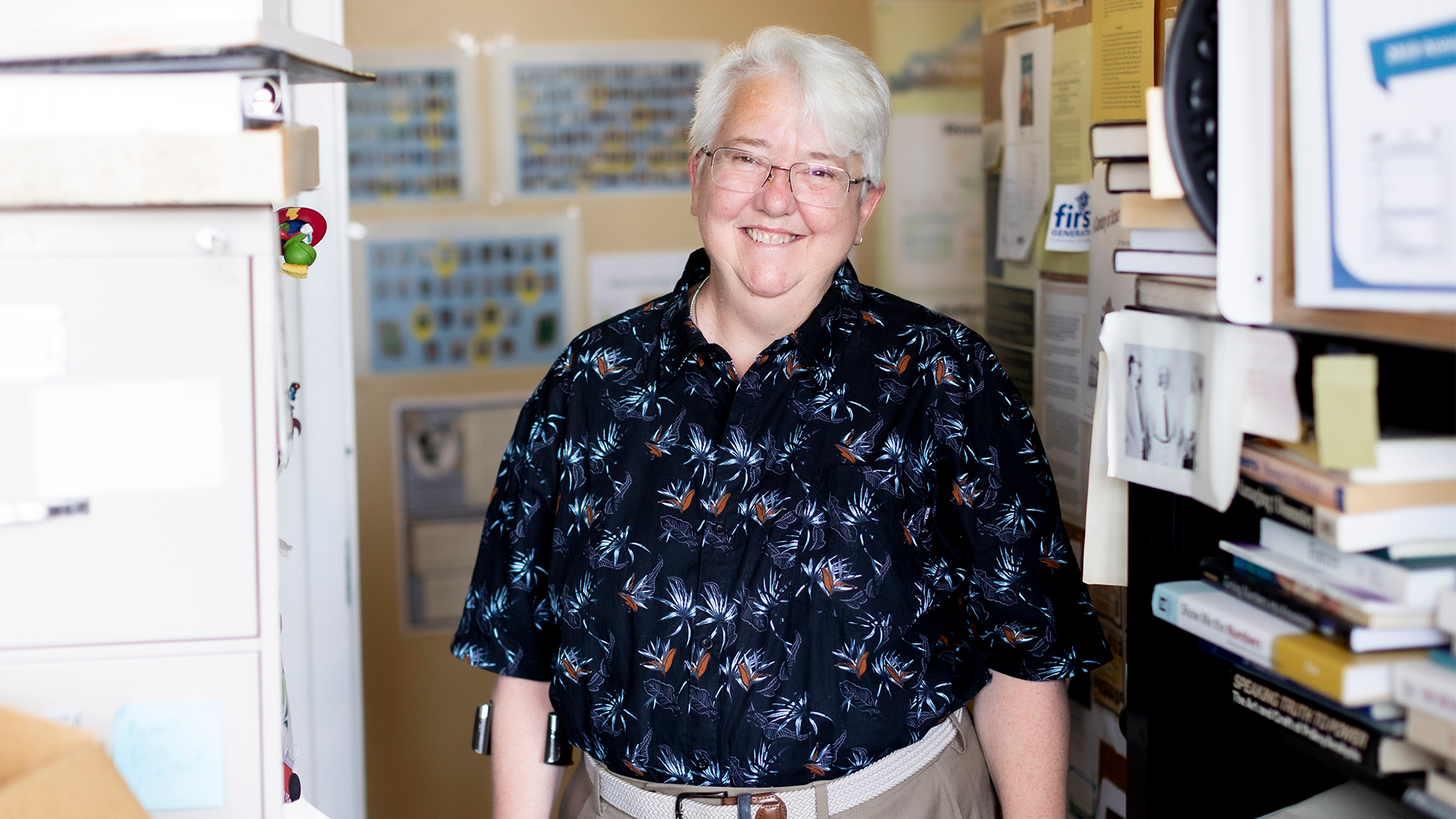A woman with short white hair, wearing a dark patterned button-up, poses for a photo in her university office. 