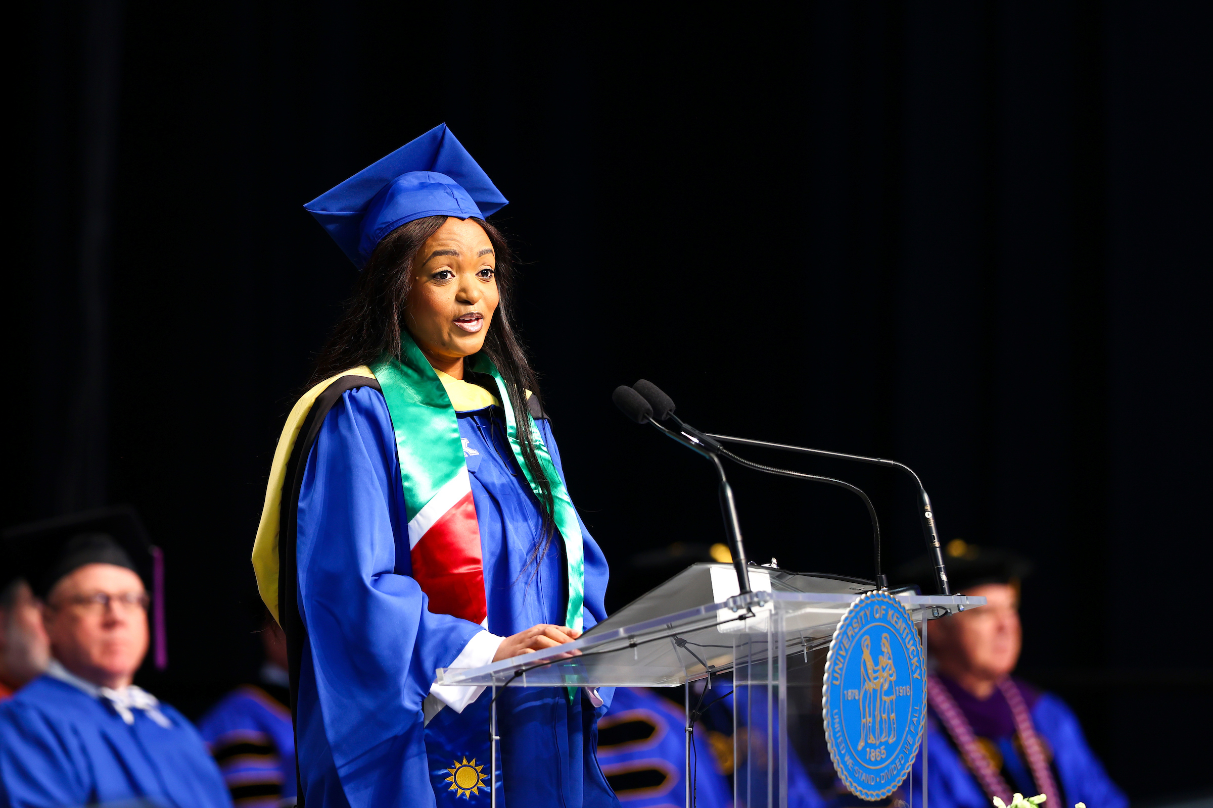 A woman in a blue cap-and-gown speaks at a podium. 