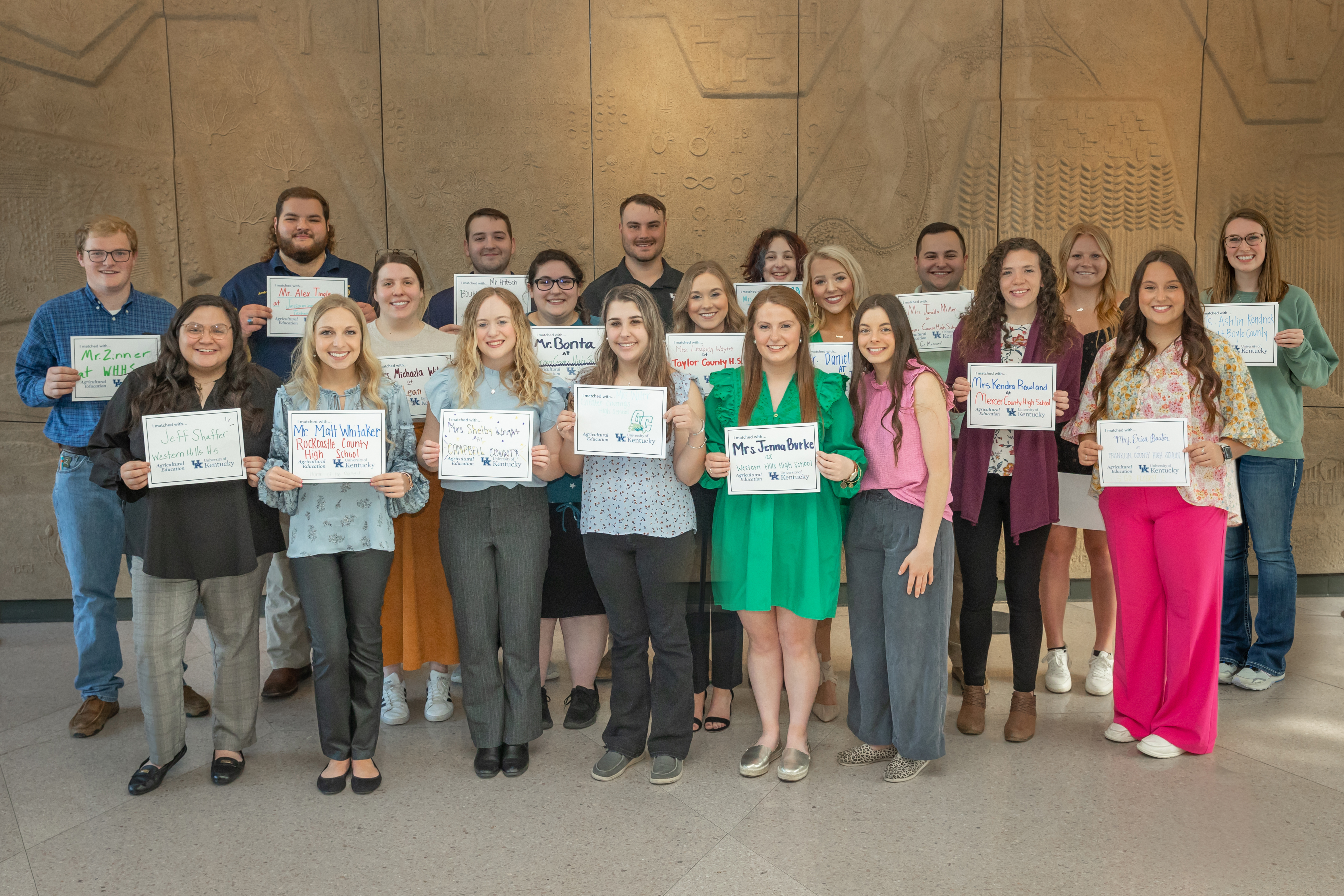 Twenty people pose together for a group photo. All are in professional dress and hold up a white piece of paper with a name or multiple names written on them.