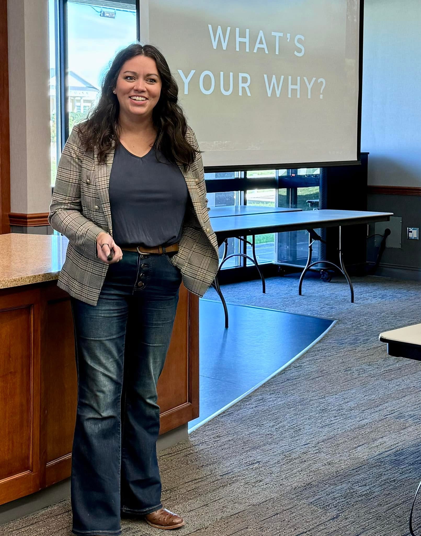 A woman in jeans and a blazer smiles while presenting at the front of a conference room. The screen to her right says "What's your why?"