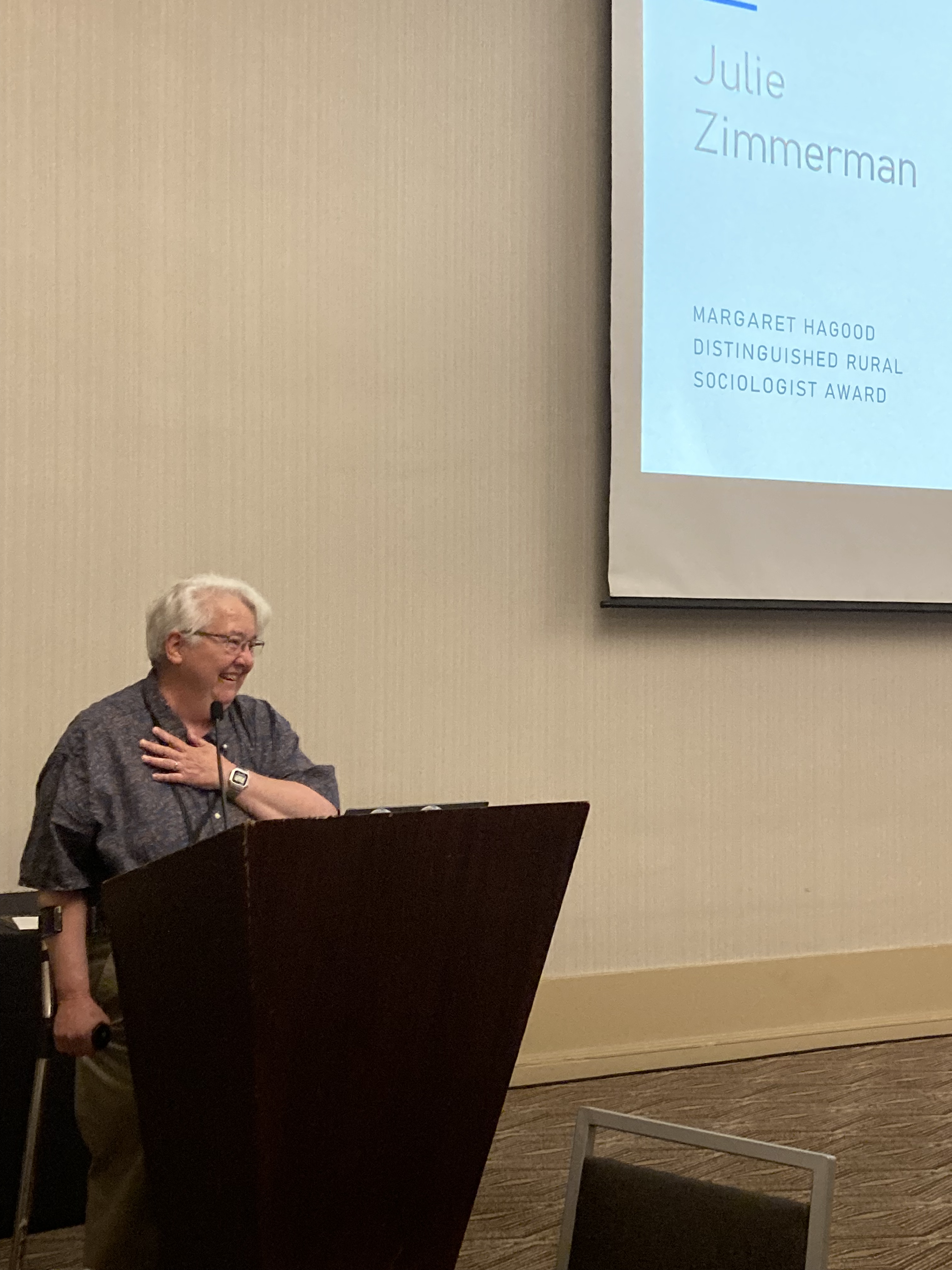 A woman with short white hair, wearing slacks and a dark-colored button-up, stands at a podium and smiles with her hand on her heart. The screen near her says her name, Julie Zimmerman.